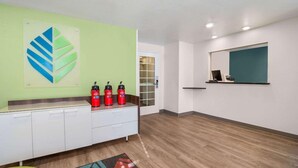 A modern hotel lobby with a light green accent wall, a large abstract artwork, three red fire extinguishers on a white counter, a teal-framed mirror, and a wooden floor. - Woodspring Suites North Charleston Airport I-526 (North Charleston)