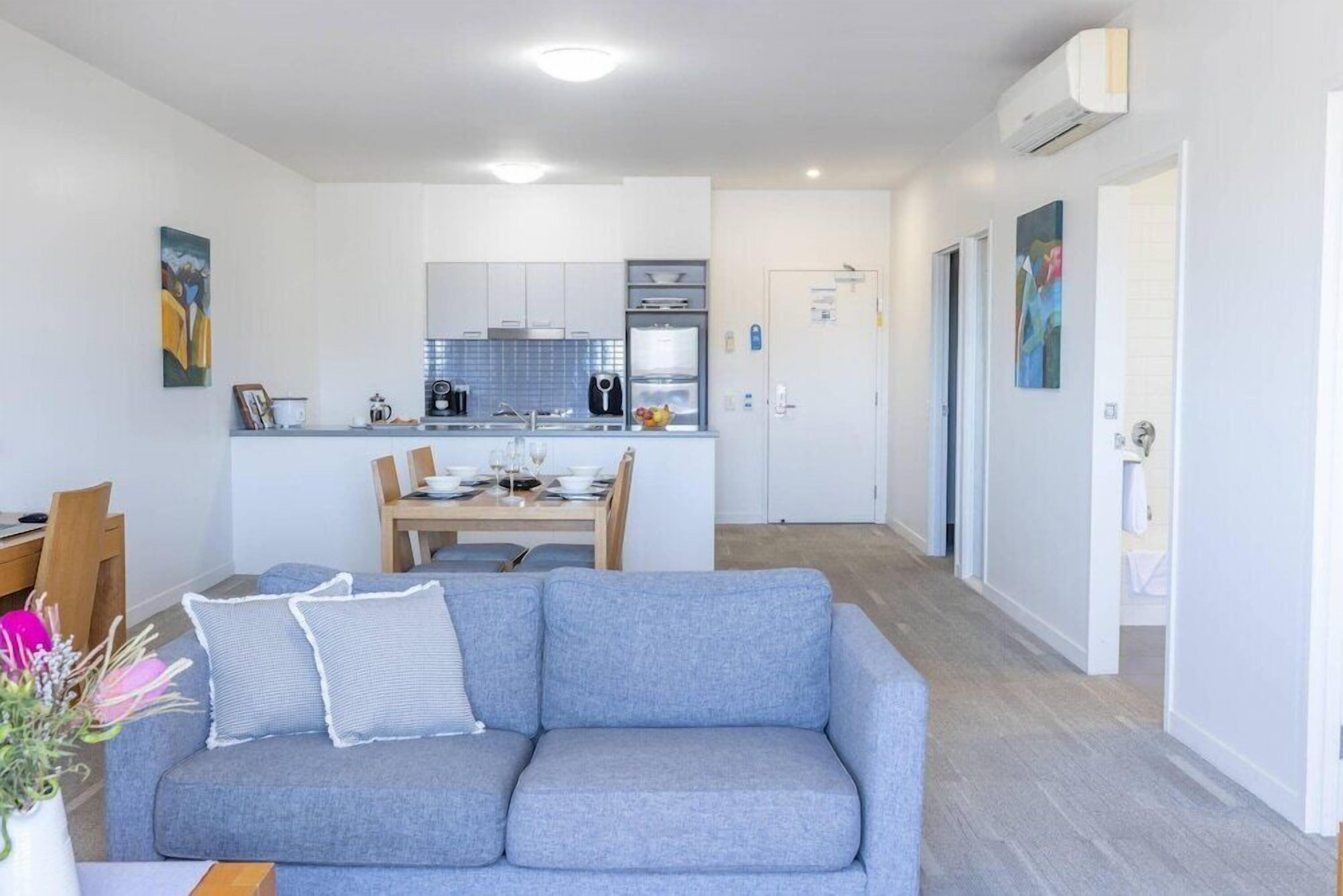 Un salon moderne avec un canapé gris, une table à manger avec des chaises et, au fond, un coin cuisine.