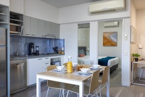 A modern kitchen with white cabinets, a blue-tiled backsplash, and stainless steel appliances. There is a dining area with a table set for two, and a bed with a blue blanket in the adjacent room. - Mainstay Suites Mackay (Mackay)