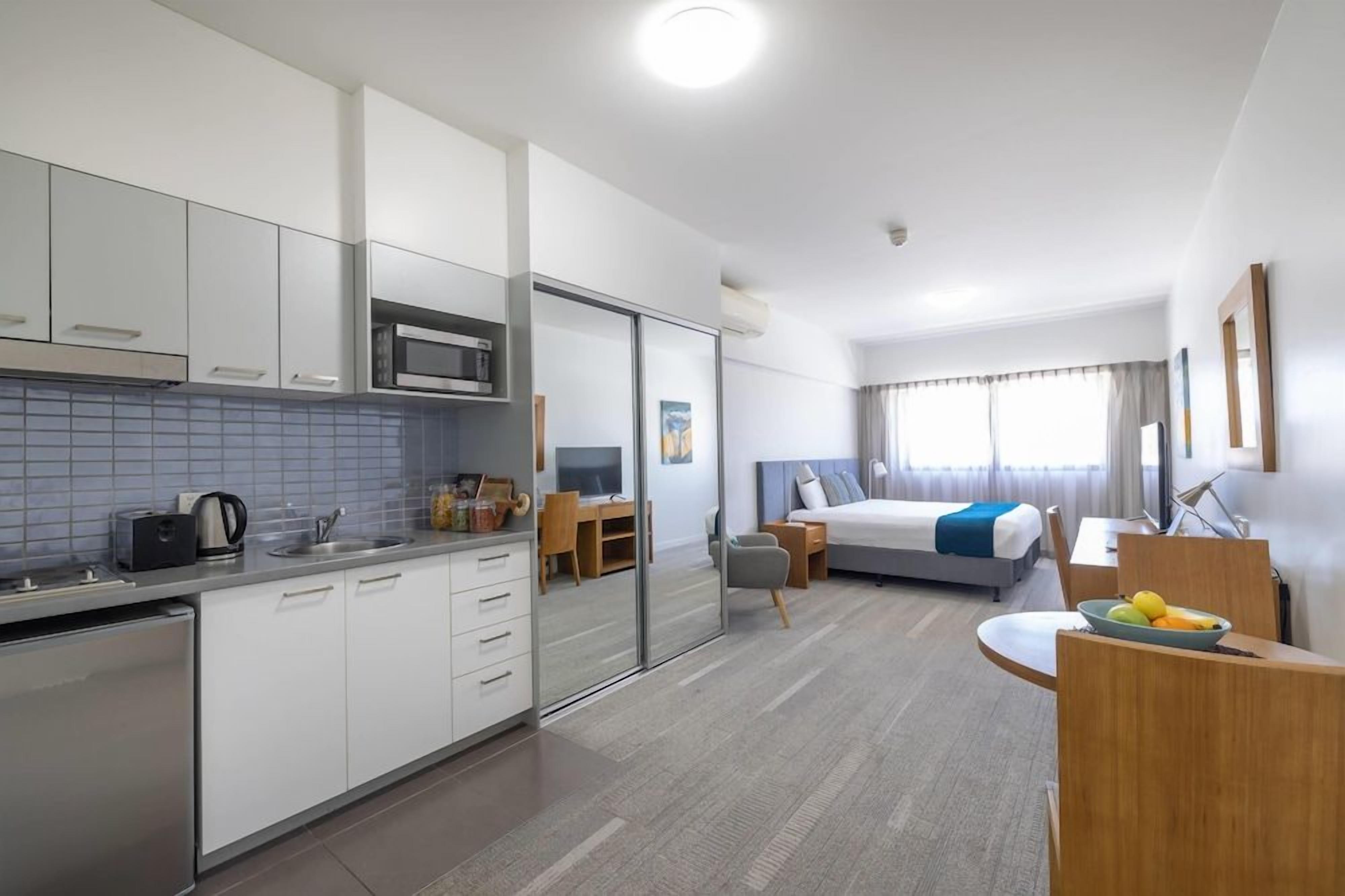 Une chambre d’hôtel moderne dotée d’une kitchenette, d’un lit aux accents bleus, d’un bureau et d’un coin repas.