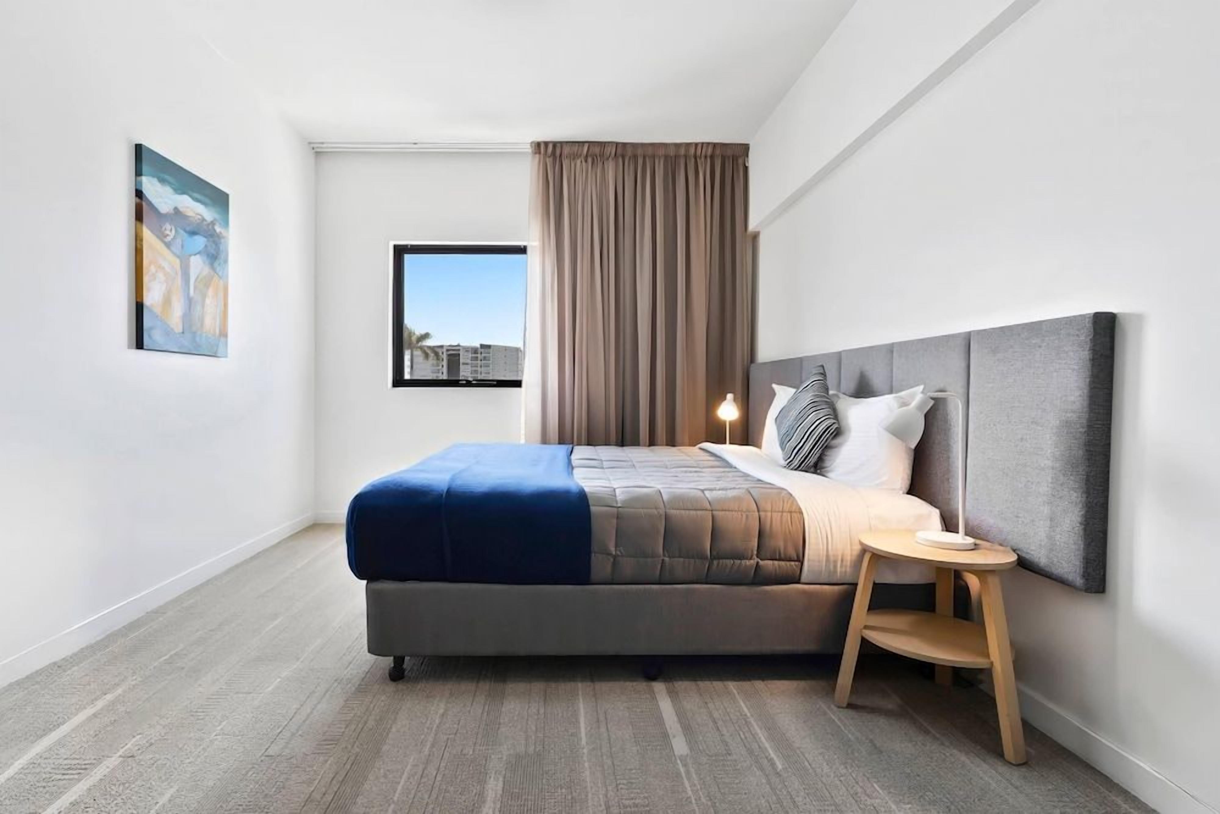 A modern hotel room with a bed, a bedside table, a window with curtains, and a painting on the wall.