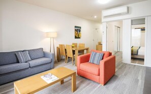 A modern living room with a grey sofa, a red armchair, and a wooden coffee table. There is a dining table with chairs and a floor lamp. An air conditioning unit is mounted on the wall. - Mainstay Suites Mackay (Mackay)