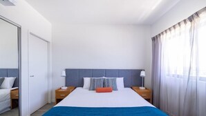 A modern hotel room with a large bed, a grey headboard, two bedside tables with lamps, a blue throw blanket, and a view of a cityscape through sheer curtains. - Mainstay Suites Mackay (Mackay)