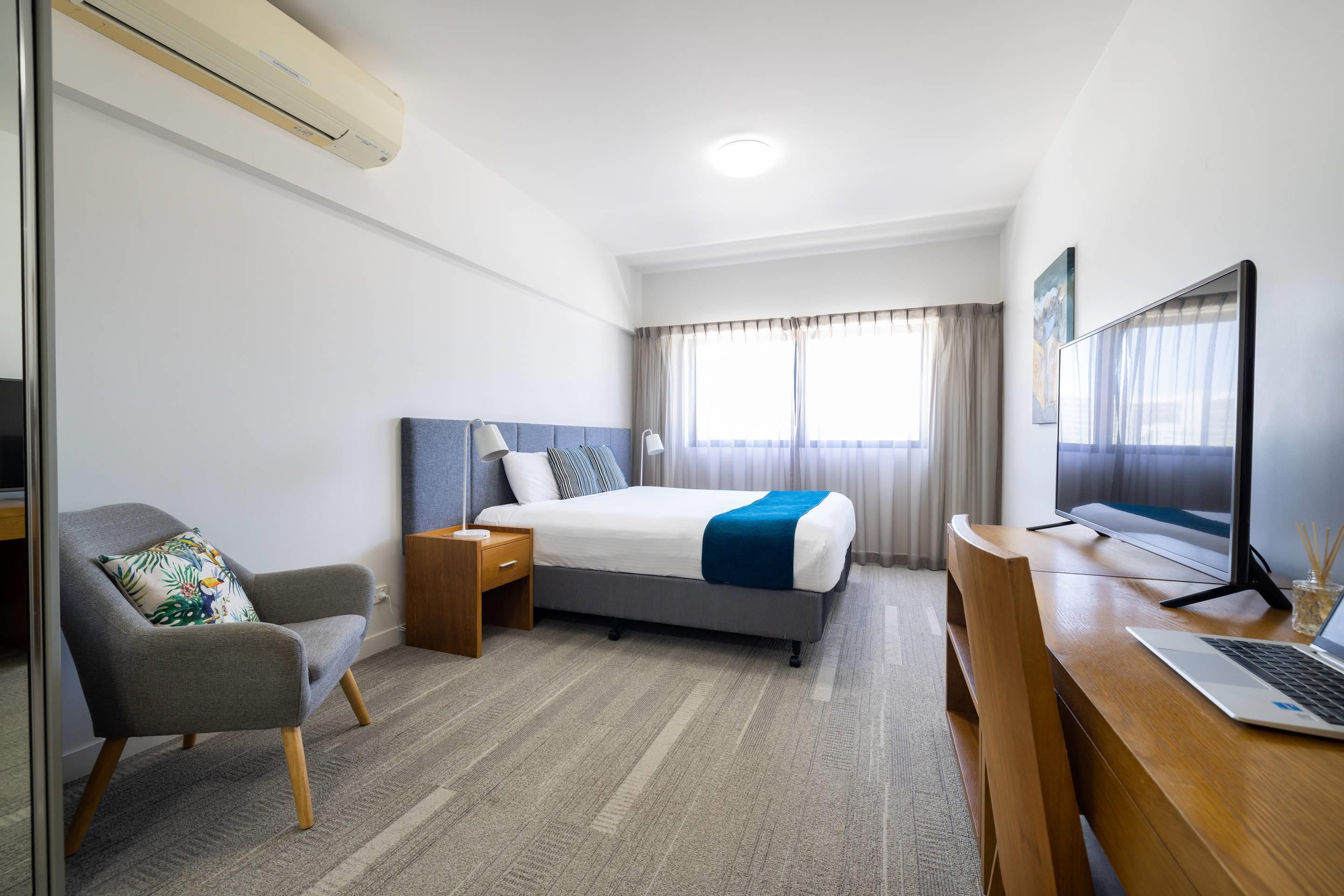 A hotel room with a bed, a desk with a laptop, a chair, a bedside table with a lamp, and a large window with curtains.