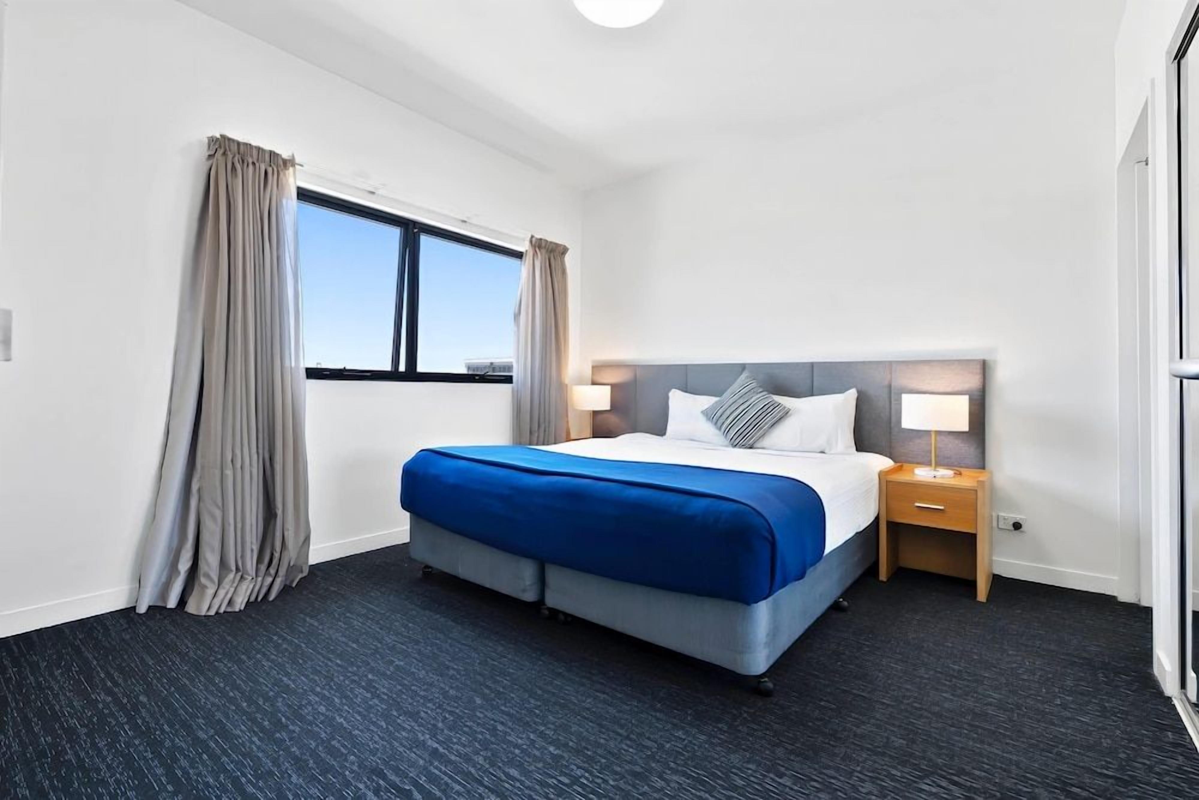 A hotel room with a bed, two bedside tables, a window with curtains, and a carpeted floor.