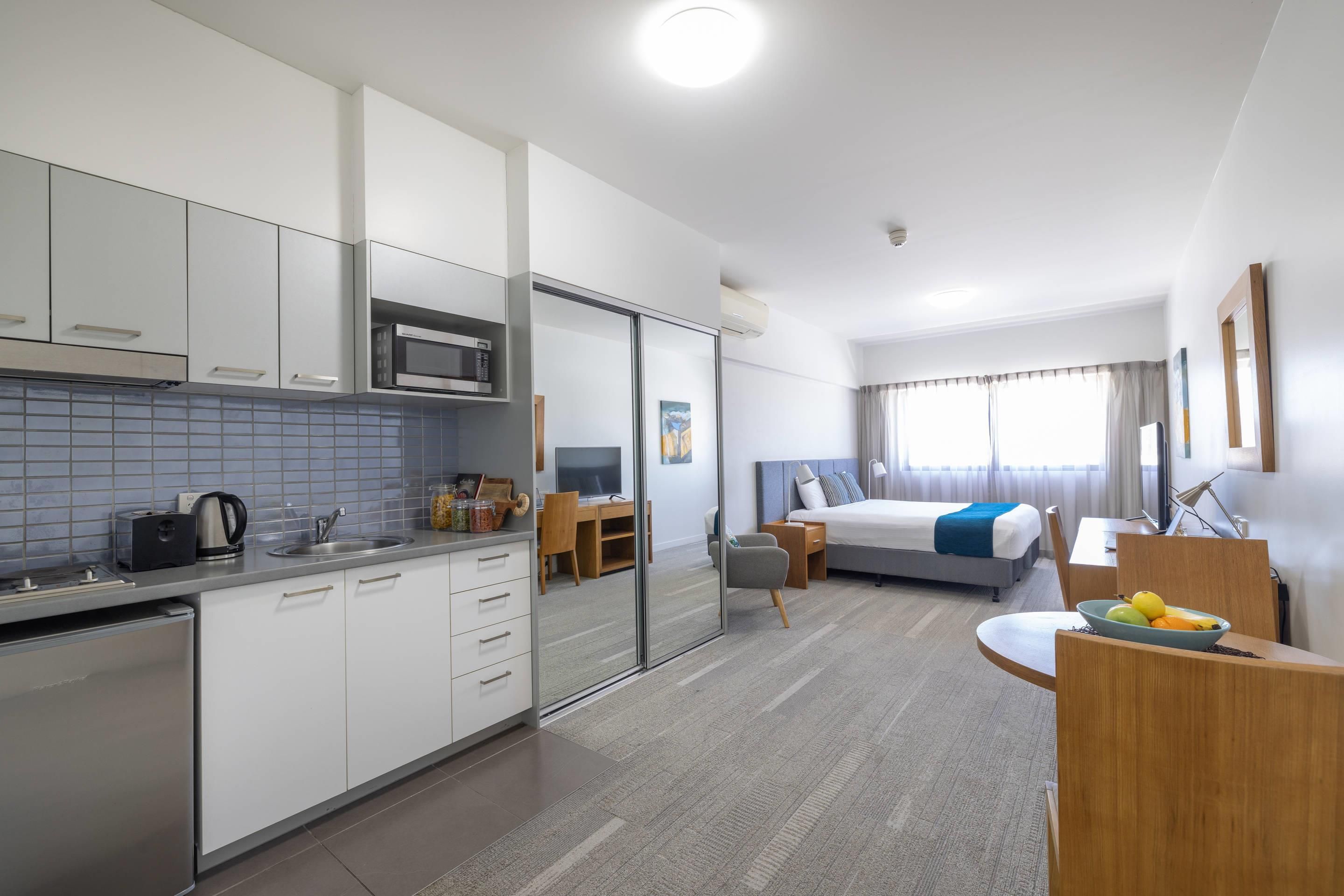 A modern hotel room with a kitchenette, a bed with a blue accent, a desk, and a large window.