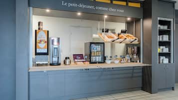 A self-service breakfast area with a coffee machine, fruit display, and a refrigerator.