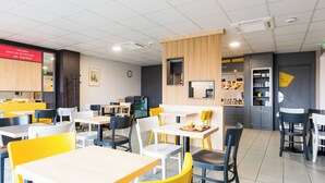 A modern dining area with multiple tables and chairs, a kitchen area in the background, and a clock on the wall. - B&B Hotel Rouen St Etienne Du Rouvray (Saint-Etienne-du-Rouvray)