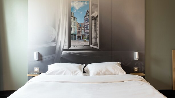 A hotel room with a bed, a nightstand, a wall-mounted lamp, and a window with a view of buildings. - B&B Hotel Rouen St Etienne Du Rouvray (Saint-Etienne-du-Rouvray)