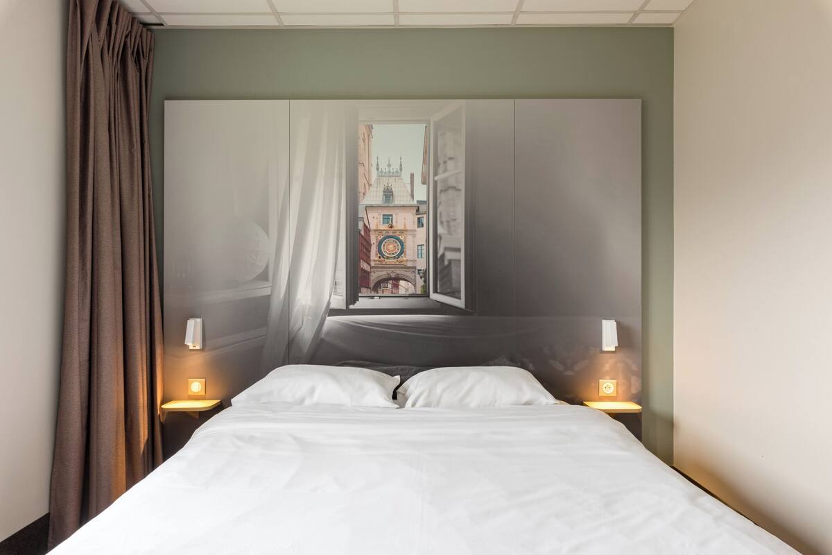 Habitación de hotel con cama, dos lámparas de noche, ventana con cortinas y vista a un edificio con torre de reloj.