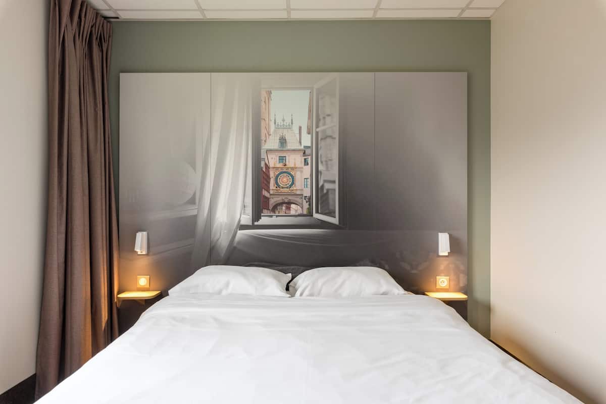 A hotel room with a bed, two bedside lamps, a window with curtains, and a view of a building with a clock tower.