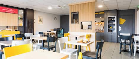 A modern dining area with multiple tables and chairs, a kitchen area in the background, and a clock on the wall.