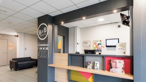 A reception area with a B&B Hotels sign, a reception desk, a computer, and a couch. - B&B Hotel Rouen St Etienne Du Rouvray (Saint-Etienne-du-Rouvray)