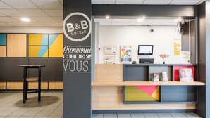 A reception desk with a B&B Hotels sign, a computer monitor, and various informational posters. - B&B Hotel Rouen St Etienne Du Rouvray (Saint-Etienne-du-Rouvray)