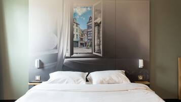 A hotel room with a bed, a nightstand, a wall-mounted lamp, and a window with a view of buildings.