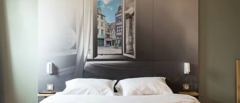 A hotel room with a bed, a nightstand, a wall-mounted lamp, and a window with a view of buildings.