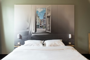 A hotel room with a bed, a nightstand, a wall-mounted lamp, and a window with a view of buildings. - B&B Hotel Rouen St Etienne Du Rouvray (Saint-Etienne-du-Rouvray)