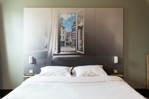 Habitación de hotel con cama, mesita de noche, lámpara de pared y ventana con vista a edificios.