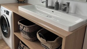 Een moderne badkamer met een wit wastafelmeubel, een spiegel, een klok en twee manden op een houten plank.