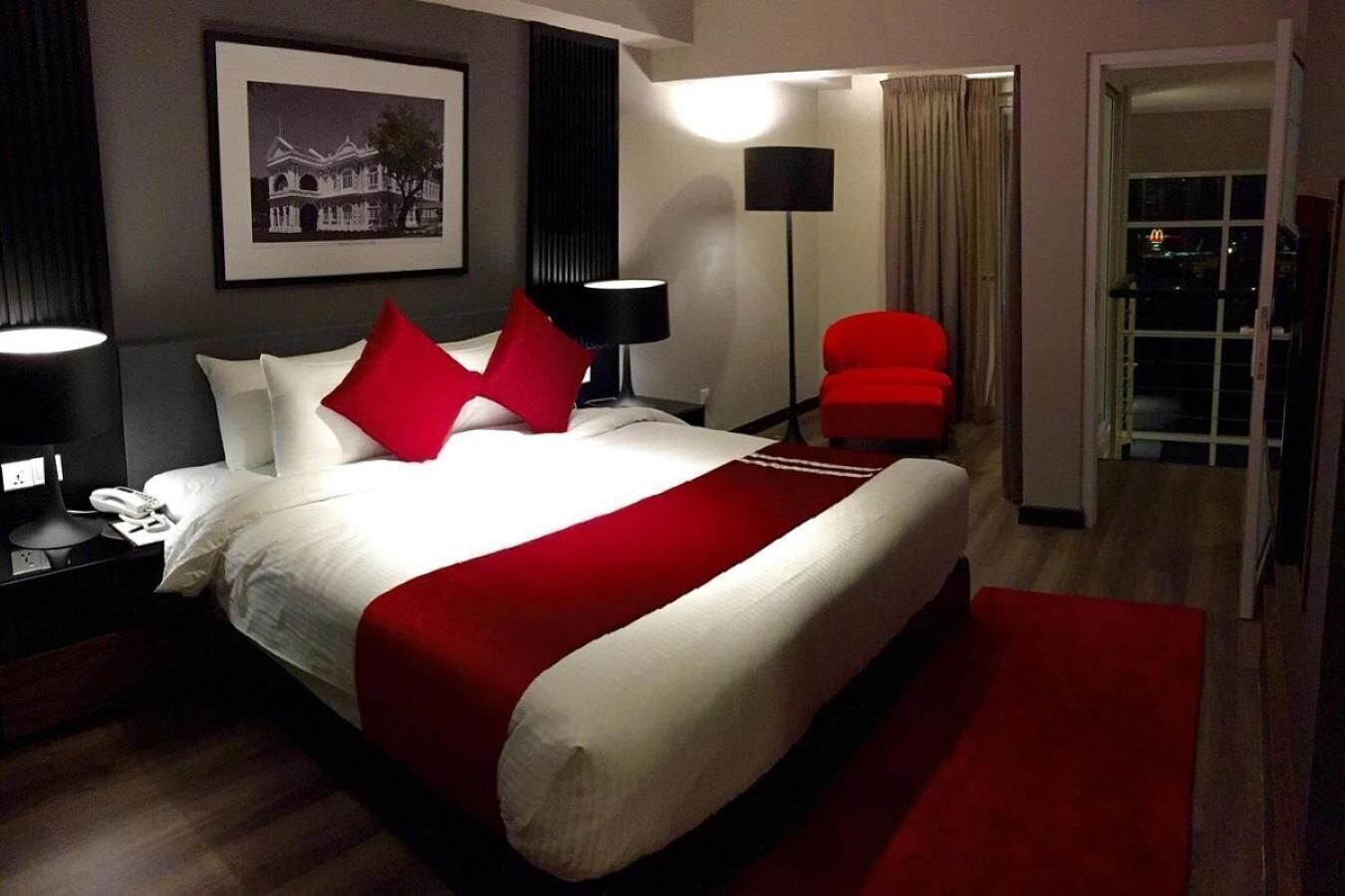 Ein Hotelzimmer mit einem großen Bett, rotem und weißem Bettzeug, einem roten Sessel, einem Nachttisch mit Telefon, einem gerahmten Bild an der Wand und einer Stehlampe.