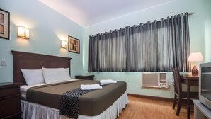 A neatly arranged hotel room with a large bed, bedside tables, a desk with a chair, a television, and an air conditioning unit. - Oftana Suites (Mandaue)