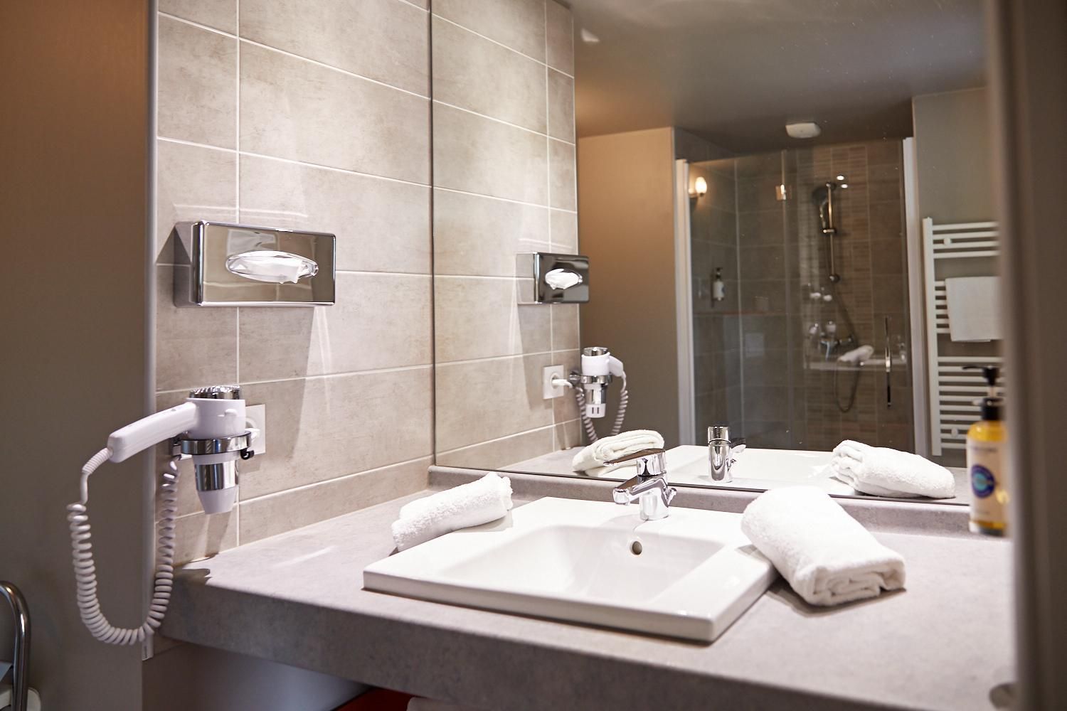 A modern bathroom with a large mirror, two sinks, a hairdryer, and a shower area.