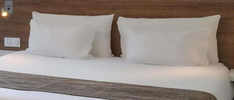 A neatly made bed with white linens and a brown throw blanket, a wooden headboard, and a wall-mounted light fixture.
