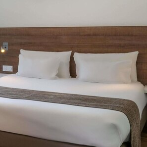 Un letto rifatto con lenzuola bianche e una coperta marrone, una testiera in legno e un lampadario fissato al muro.
