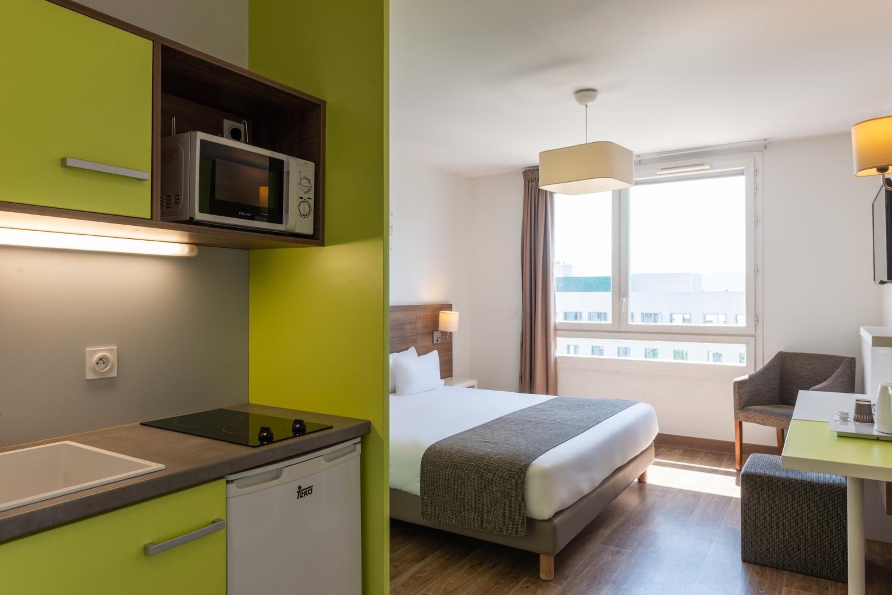 A hotel room with a bed, a desk, a chair, a window with a view, a microwave, and a small kitchenette.