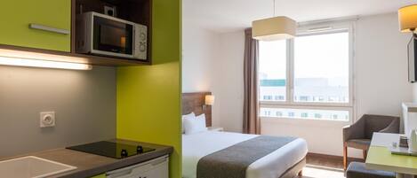 A hotel room with a bed, a desk, a chair, a window with a view, a microwave, and a small kitchenette.