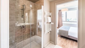Ein modernes Hotelzimmer mit einer gläsernen Duschkabine, einem Bett mit grauer Decke und Blick ins Freie durch ein großes Fenster.