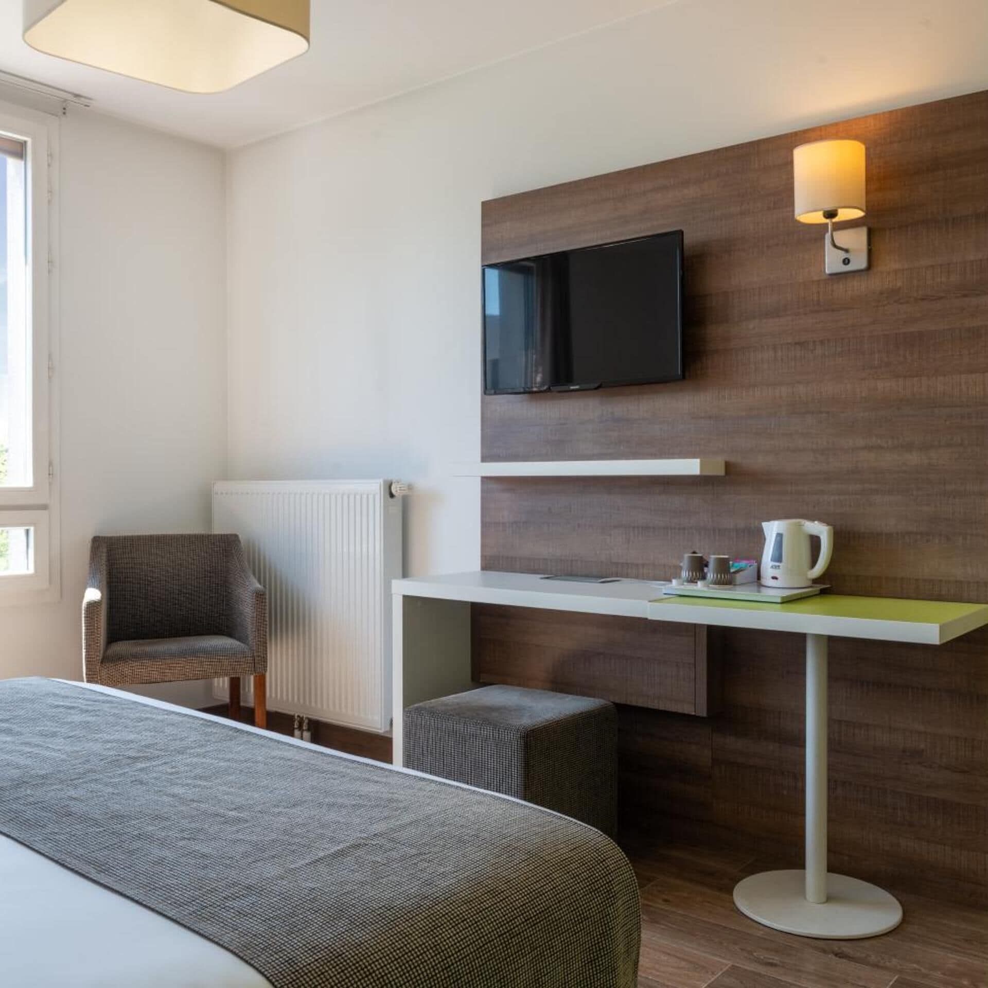 A hotel room with a bed, a chair, a desk with a television, a kettle, and a radiator.