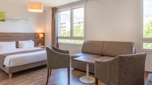 Ein Hotelzimmer mit einem Bett, zwei Stühlen, einem Sofa, einem Tisch und großen Fenstern mit Blick auf Gebäude und Bäume.