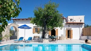 A pool area with a clear blue pool, lounge chairs, and a tree in the center. - Silver Gem Villa with Private Pool (Frantzeskiana Metochia)