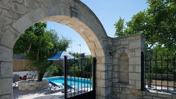 A stone archway leading to a swimming pool area with lounge chairs and a blue umbrella. - Silver Gem Villa with Private Pool (Frantzeskiana Metochia)