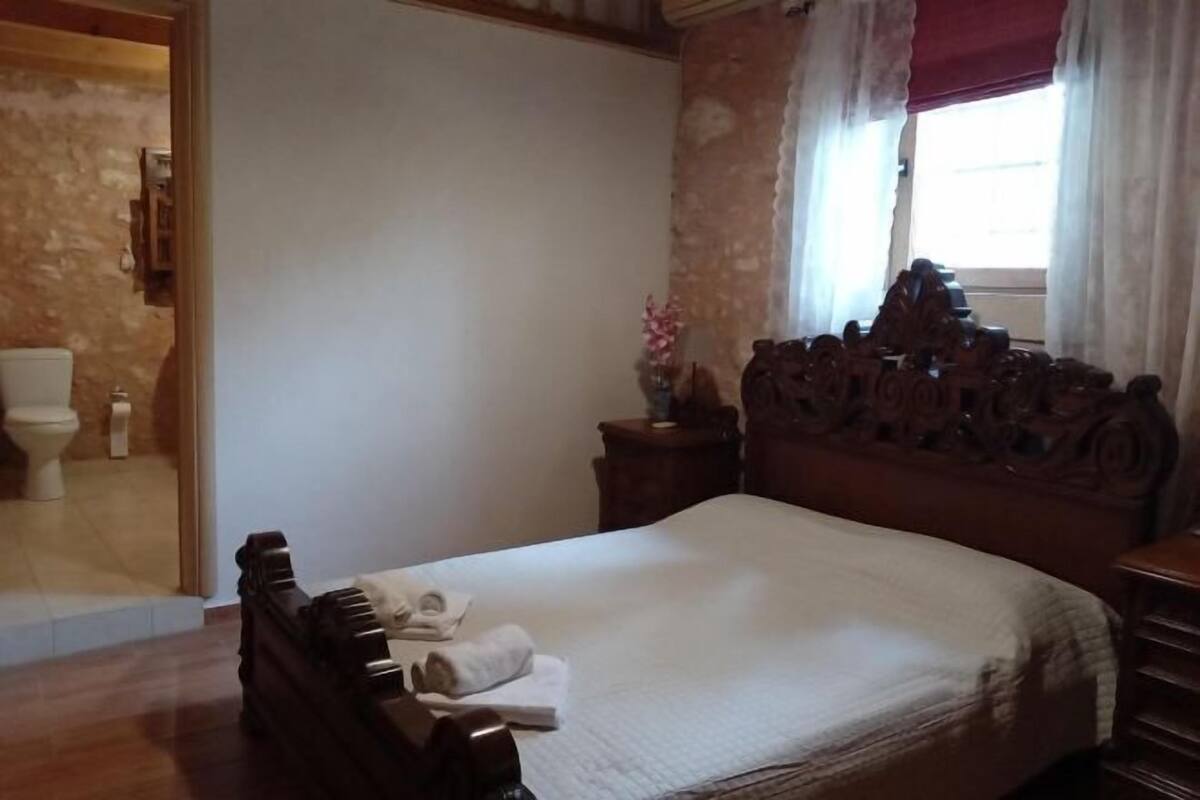 A bedroom with a wooden bed, a nightstand with a vase, and a bathroom visible through an open door.