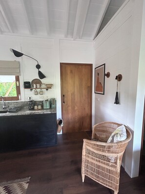 Una stanza con pavimento in legno scuro, una sedia in vimini, un angolo cottura con un piano di lavoro e una porta in legno.