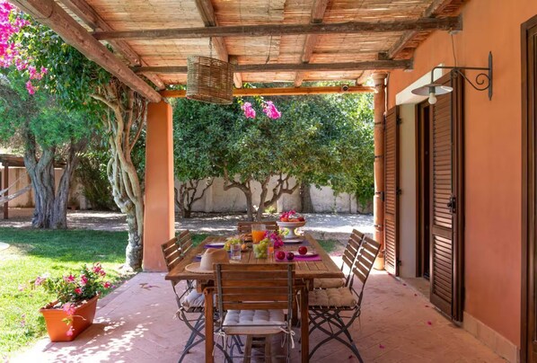 A covered outdoor dining area with a table set for two, surrounded by chairs, and a potted plant.