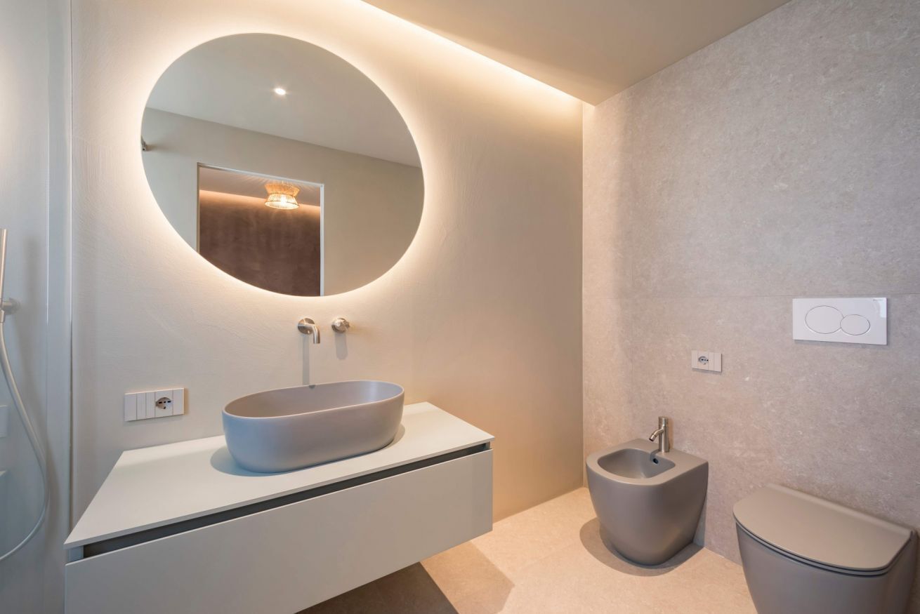 A modern bathroom with a round mirror, a vessel sink, a bidet, and a toilet.