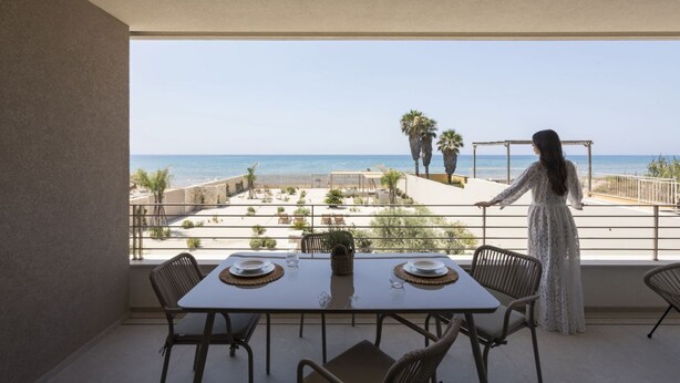 A balcony with a table set for two, overlooking a beach with palm trees and a clear sky. - Cala Theresia, Alea - Sea front apartment (Pachino)