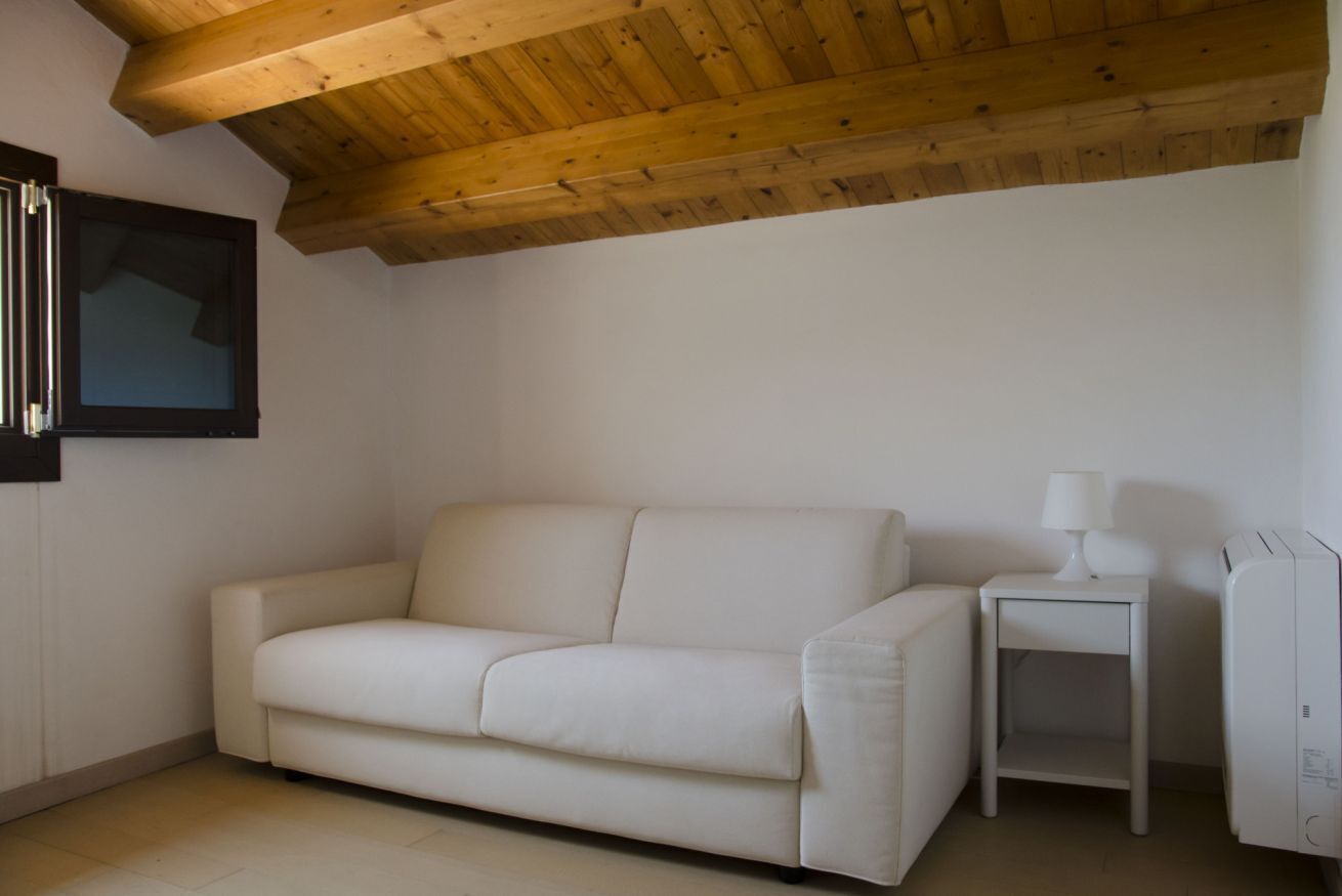 A room with a wooden ceiling, a white sofa, a small white table with a lamp, and a white cabinet.