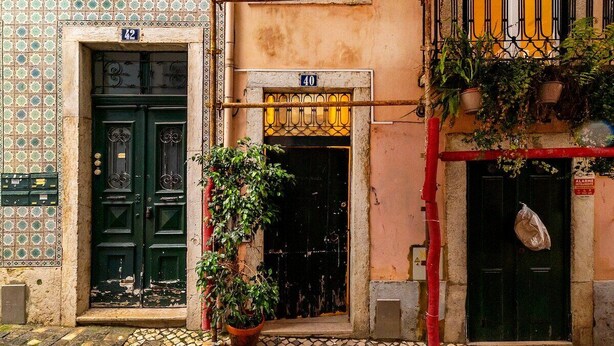 Um edifício com duas portas, uma marcada com o número 42 e a outra com 40. Há uma planta em vaso em frente às portas, e um cano vermelho percorre a parede.