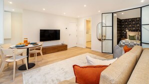 Uma sala de estar moderna com um sofá, uma mesa de jantar com cadeiras, uma televisão de tela plana e uma cozinha visível através de um painel de vidro.