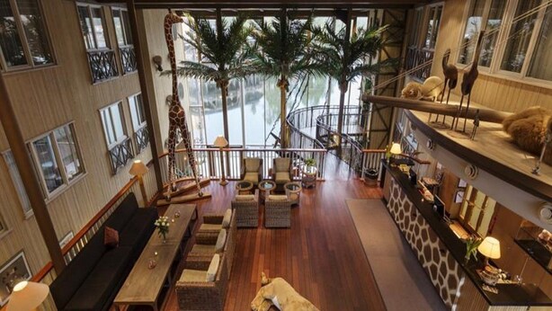 A spacious living area with a wooden floor, a sofa, a dining table, and a reception desk. There are large windows with a view of a body of water and palm trees. - Dyreparken Safarihotell (Kristiansand)