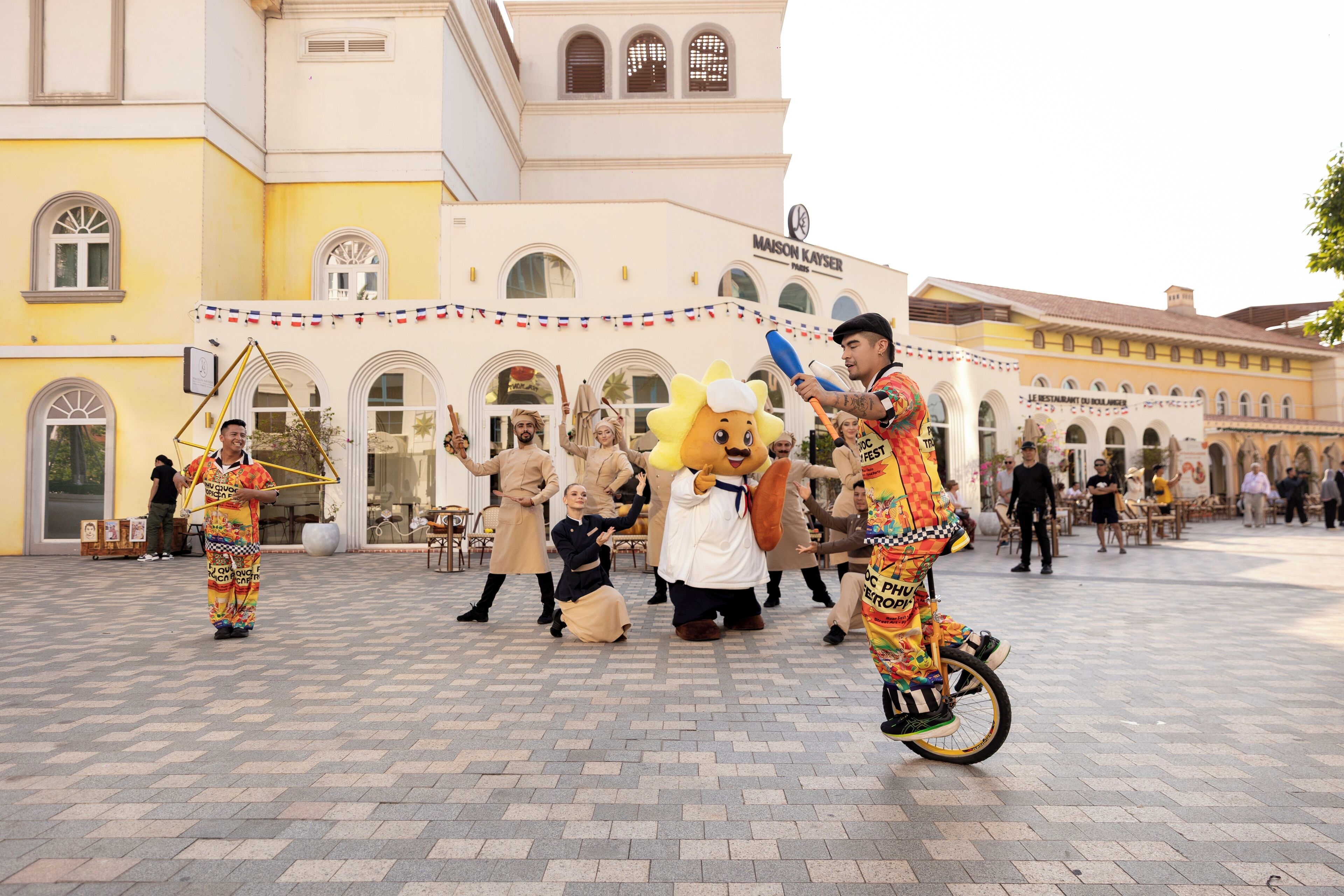 A group of performers in colorful costumes dancing in a plaza with a building labeled "MAISON KAYSER" in the background.