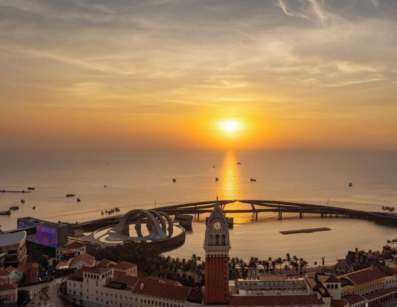 Une vue urbaine côtière avec une tour de l’horloge imposante, un pont et une structure circulaire au bord de l’eau, le tout baigné par les couleurs du coucher de soleil.