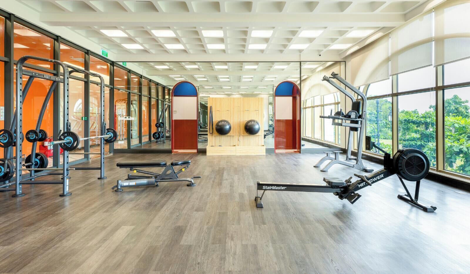A gym with various exercise equipment, large windows, and a wooden floor.