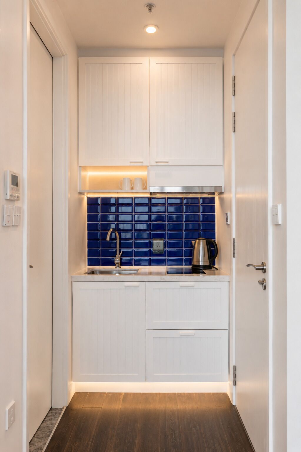 A narrow hallway leading to a compact kitchen with white cabinets, a blue backsplash, and a coffee maker on the counter.