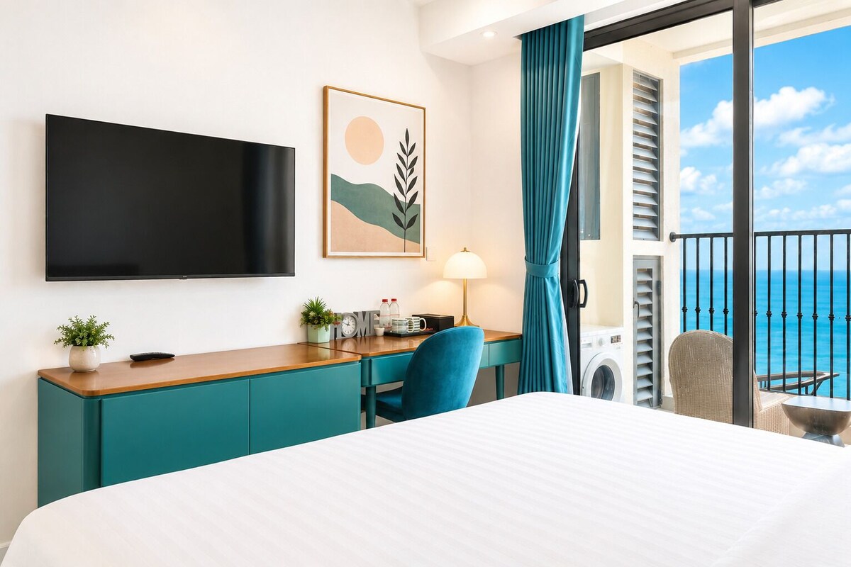 A hotel room with a bed, a desk with a lamp, a TV, a view of the ocean, and a balcony with a chair.