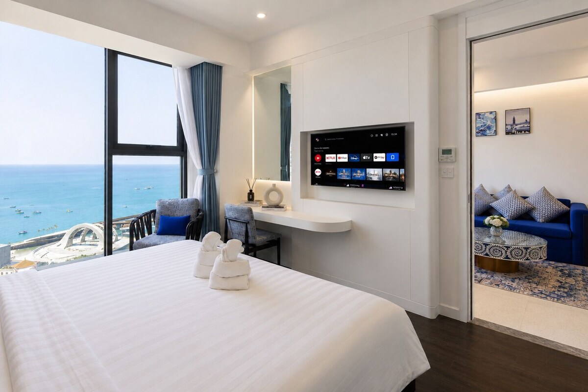 A hotel room with a large bed, a TV mounted on the wall, a seating area with a sofa and chairs, and a view of the sea.